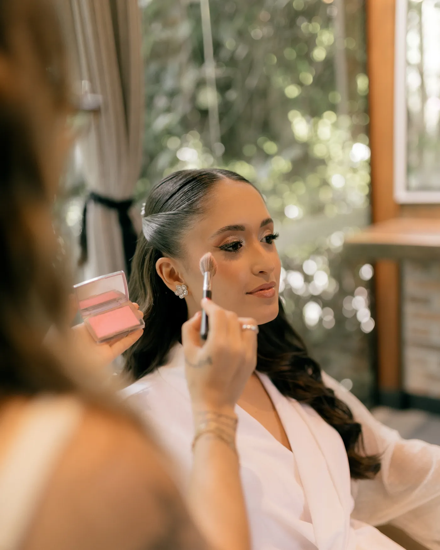 Makeup artist applying blush to a woman in a white robe while she gets ready
